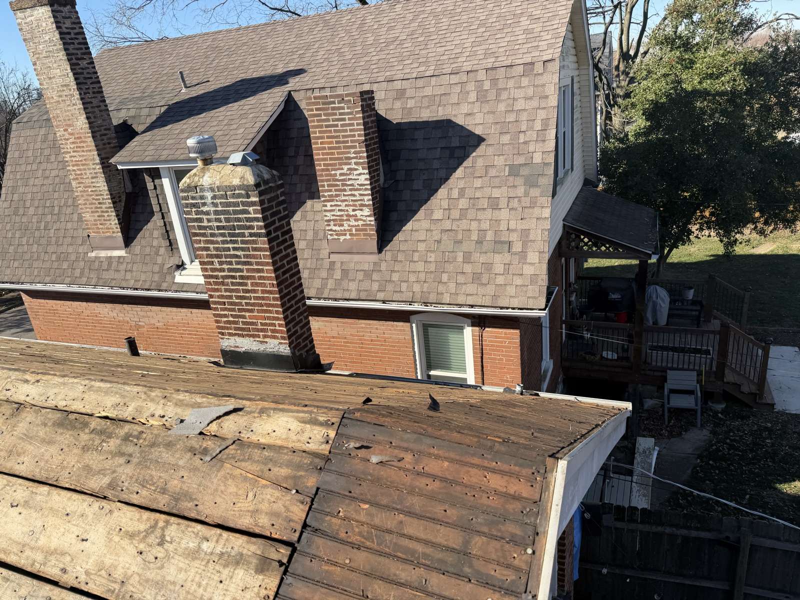 A lower roof stripped to plank decking in the foreground with felt scraps and old nails, and behind it a two storey red brick house still carrying weathered brown shingles, with two brick chimneys, a chimney breast running up the wall whose brick faces have spalled leaving pale patches, brown metal flashing at the junctions where the small roof returns meet the brick, a dormer with a metal turbine vent, and a raised wooden deck to the right