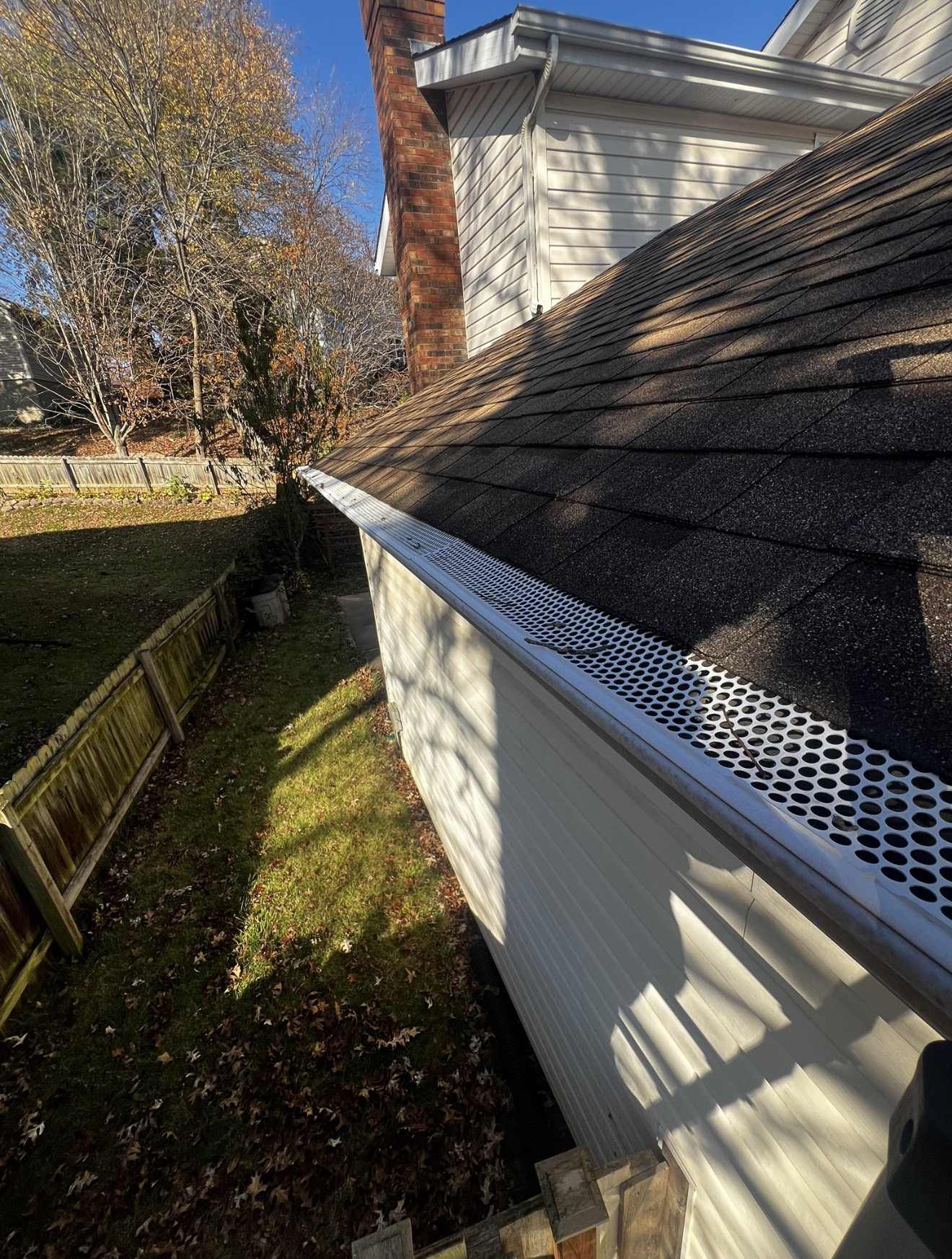 Along the eave of a dark grey brown shingle roof in strong low sun, with a white perforated metal gutter guard punched with round holes running the length of the gutter and two bare twigs and a few small dark scraps of debris lying on top of the perforations, a red brick chimney and white vinyl siding above, a wooden privacy fence and a back lawn with fallen autumn leaves below