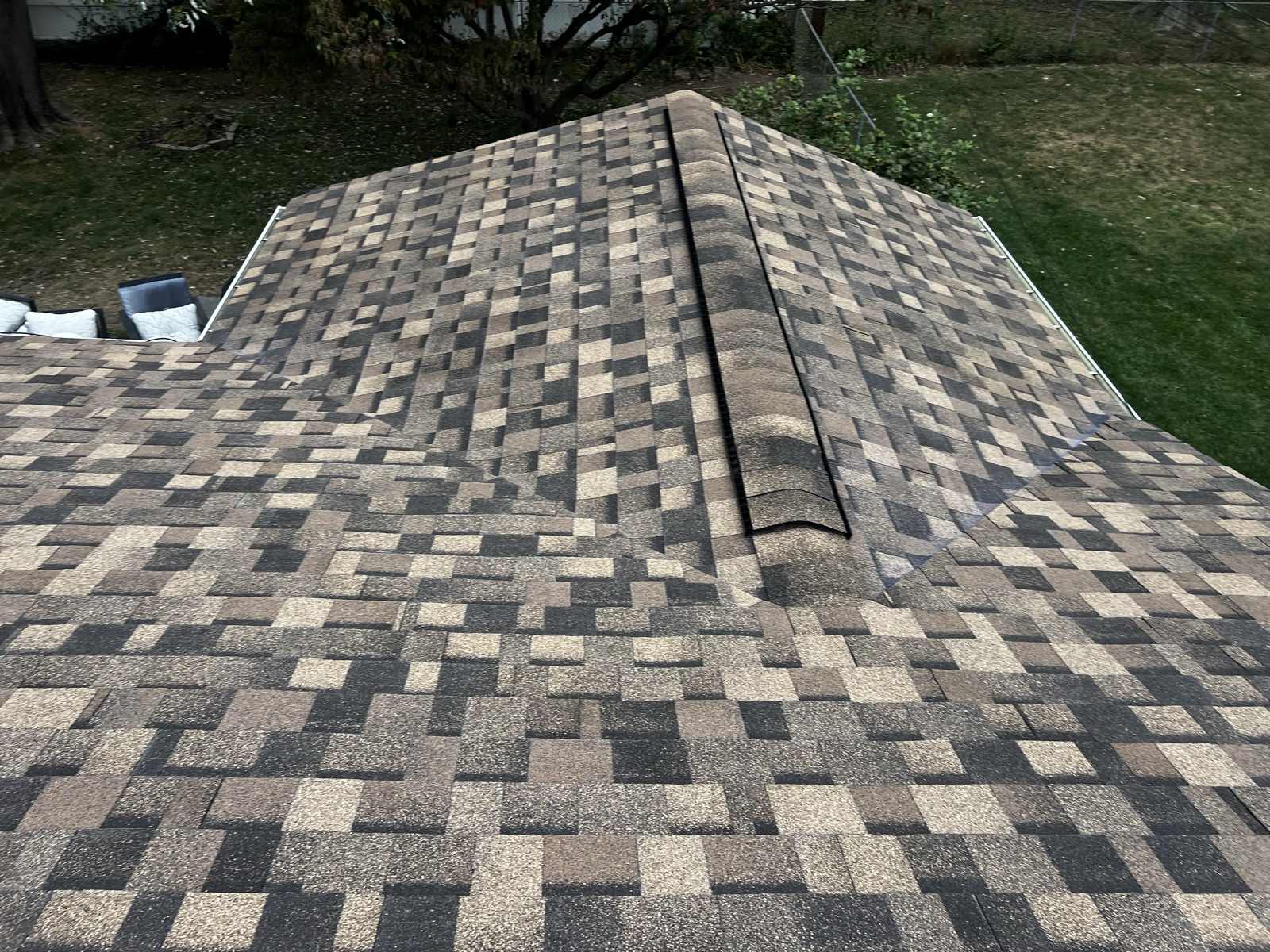 Looking down at the hip of a brown, tan and charcoal multi tone architectural shingle roof, capped along the hip with ridge cap shingles and carrying a dark vent strip beside the cap, with gutters running along the eaves either side and a mown lawn, chain link fence and patio furniture in the yard below