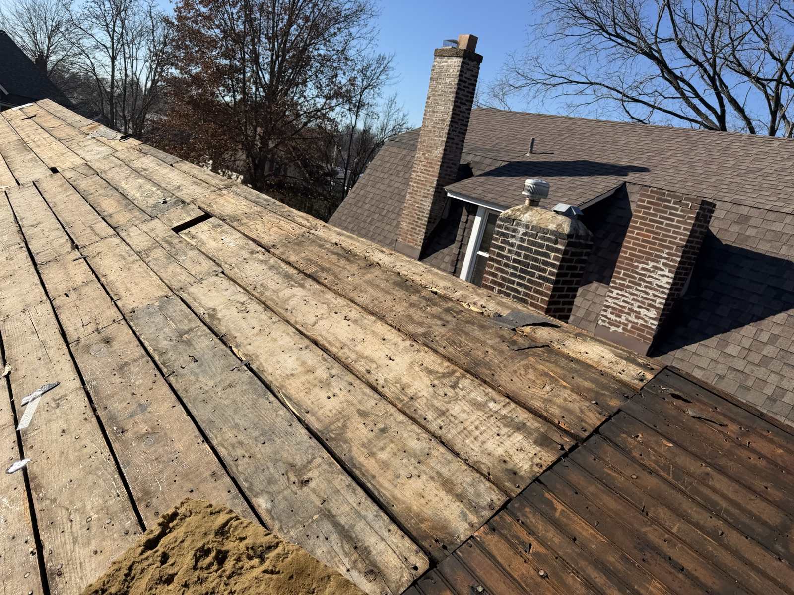 A roof plane stripped back to wide wooden plank decking with visible gaps between the boards and old nails still standing proud, a sawdust and granule pile at the lower left, and beyond it the main house still carrying weathered brown shingles with two brick chimneys showing spalling and patched mortar and a dormer with a metal turbine vent, under a clear winter sky
