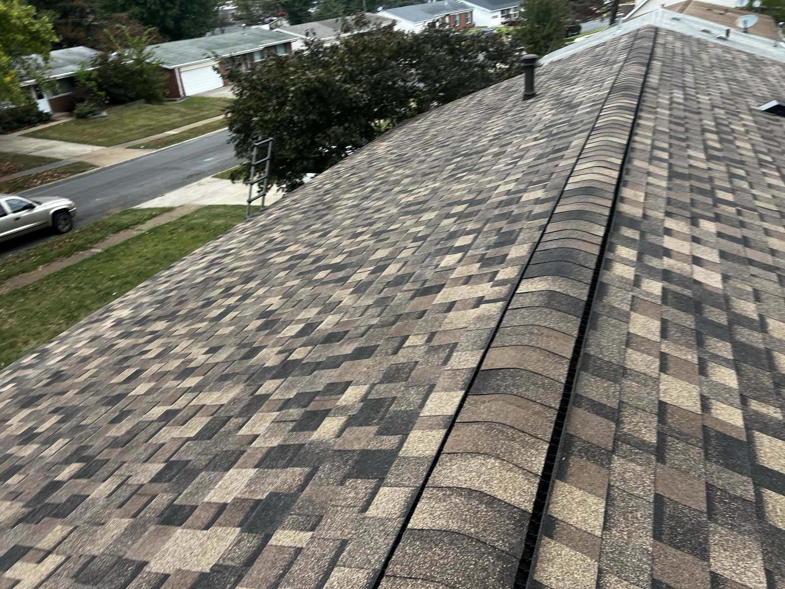 Brown, tan and charcoal multi tone architectural shingles running up to a capped ridge with a dark ridge vent slot beneath the cap and a metal flue cap near the far end, an aluminium ladder standing at the eave in the upper left, and a suburban street of single storey brick and vinyl ranch houses with a white pickup at the kerb below