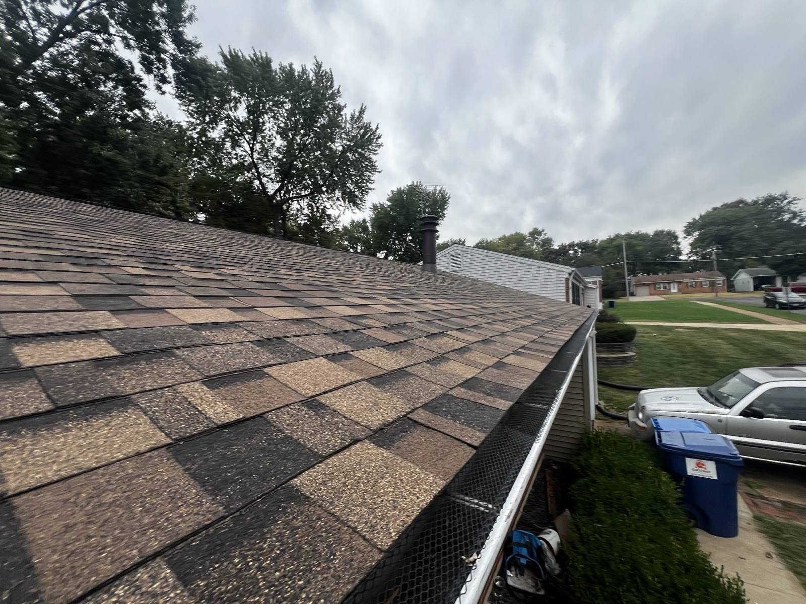 Worn brown and tan shingles along an eave seen from the roof surface, with pale patches of granule loss across the field, a black mesh gutter guard over the white gutter, a metal flue cap at the ridge line and a suburban street with a driveway, parked vehicles and a recycling bin below